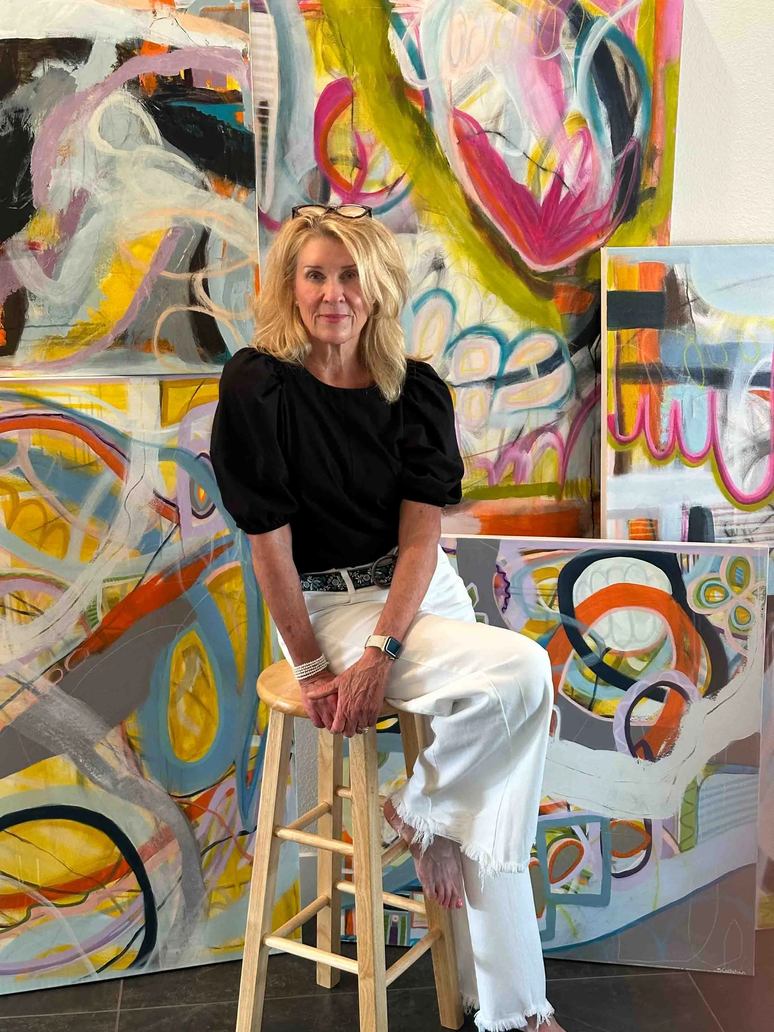 A woman with blonde hair, wearing a black top and white pants, sitting on a stool in front of colorful abstract paintings, with glasses on her head and a smartwatch on her wrist.