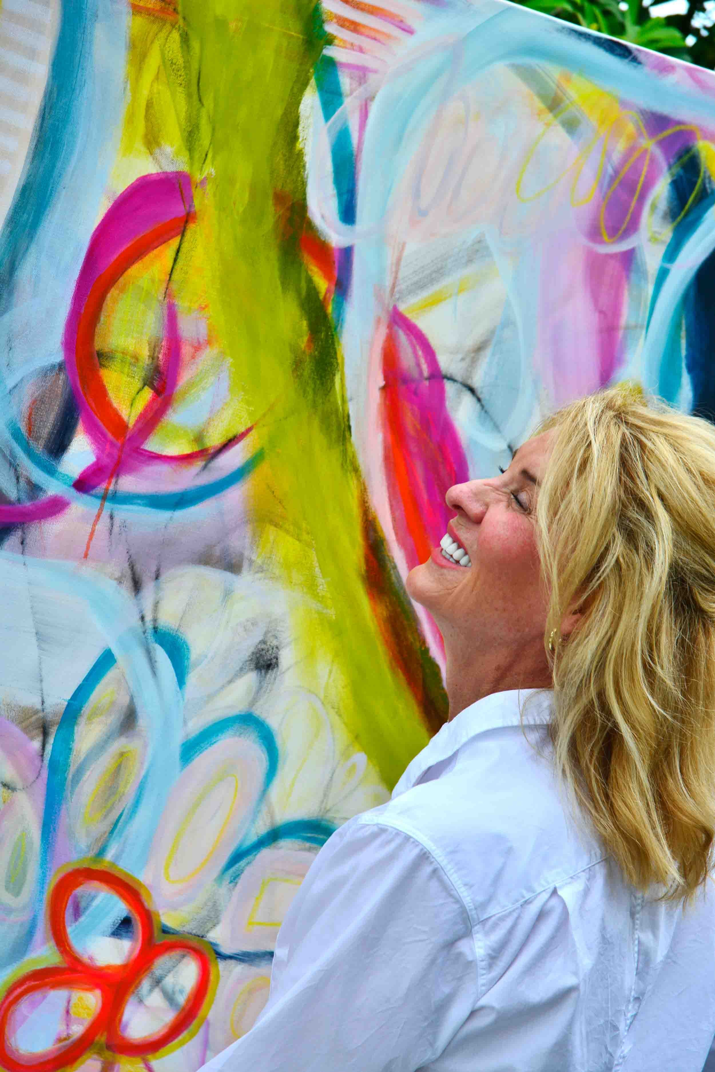 A woman with blonde hair smiling happily as she looks at a colorful abstract painting filled with vibrant swirls and circles in shades of pink, blue, yellow, and orange.