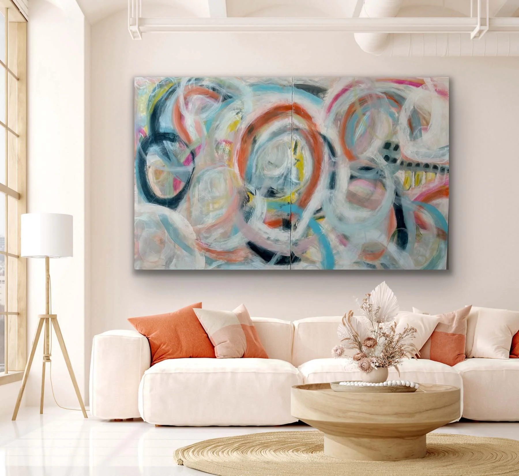 Modern living room with white sofa, orange and white pillows, abstract colorful painting on the wall, wooden coffee table with floral decor, standing lamp, and natural lighting.