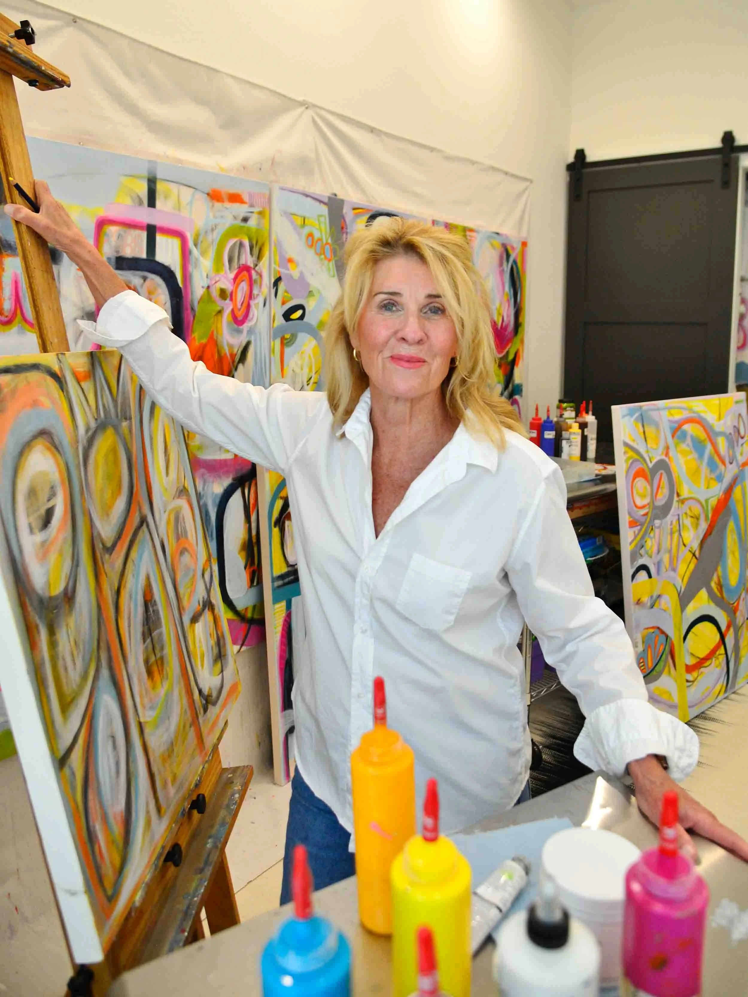 An older woman with blonde hair standing in front of colorful abstract paintings in an art studio, with bottles of brightly colored paint and brushes on a table in the foreground.