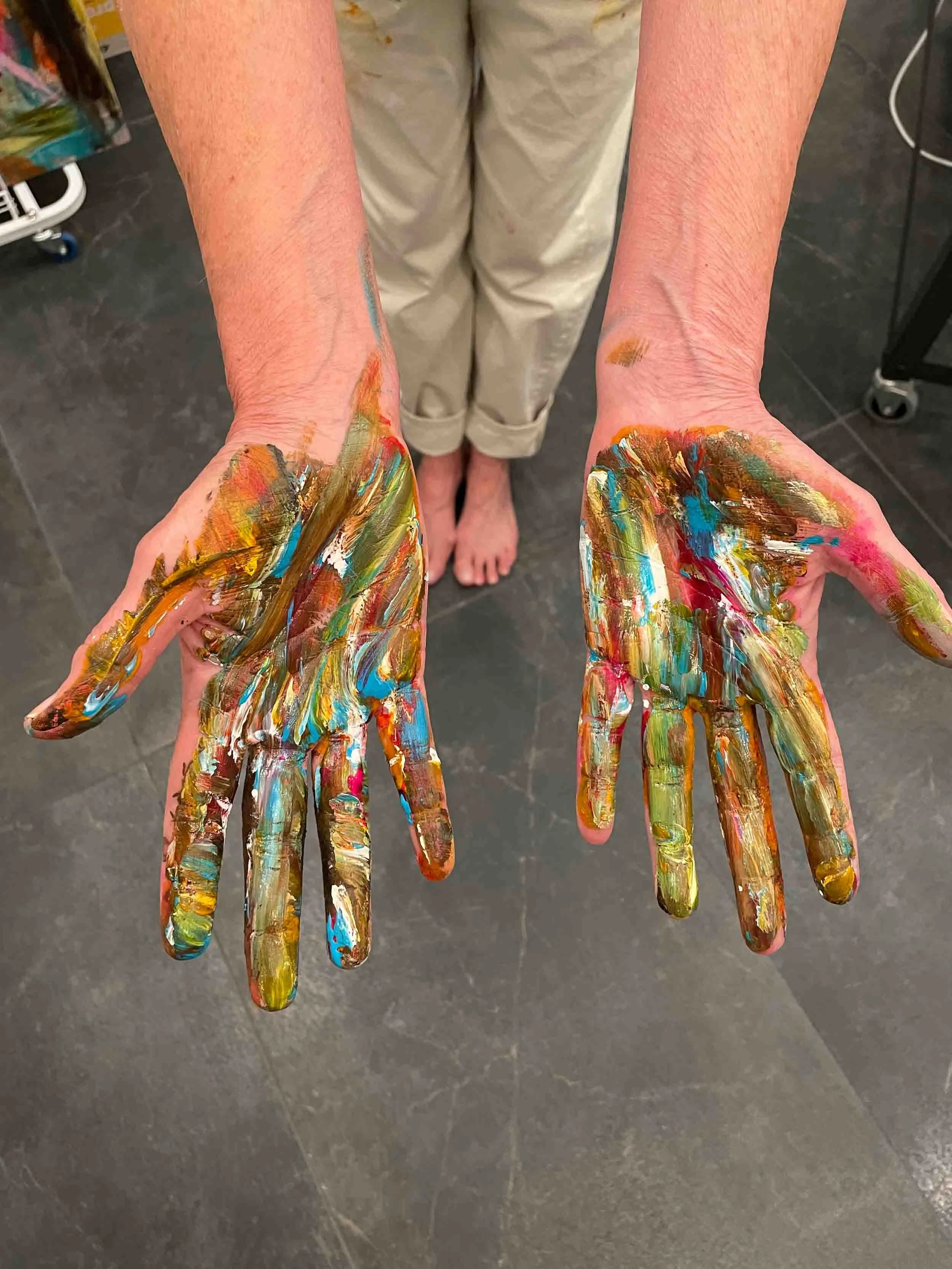 Hands covered in colorful paint, with a person standing barefoot in the background on a dark floor.