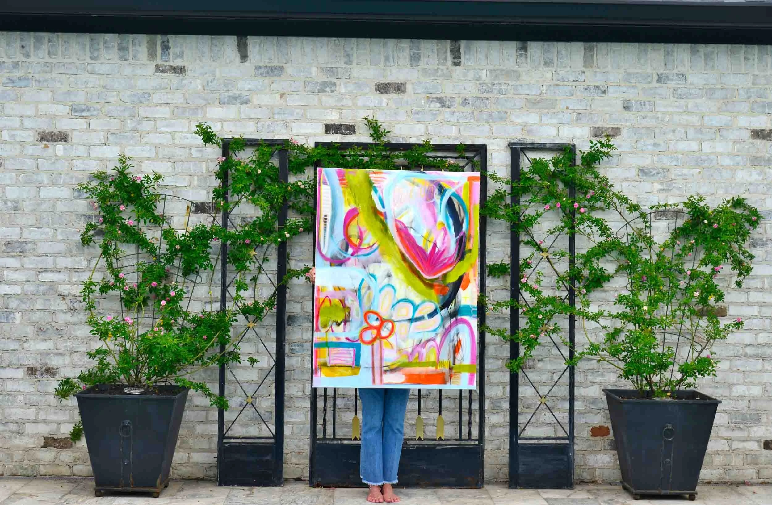 Person standing in front of brick wall decorated with potted plants and black metal frames, holding a colorful abstract painting that covers their face.