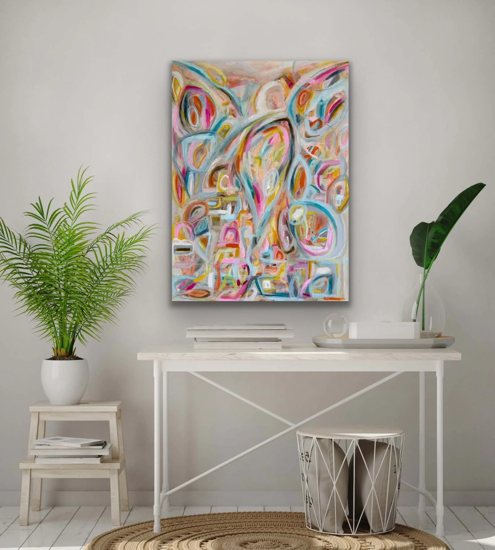 Interior scene with a colorful abstract painting on the wall above a white table. The table has a stack of books, a glass globe, and a decorative tray with a green plant in a tall vase. To the left, there is a small wooden stool with more books, and on the floor, a round woven rug.