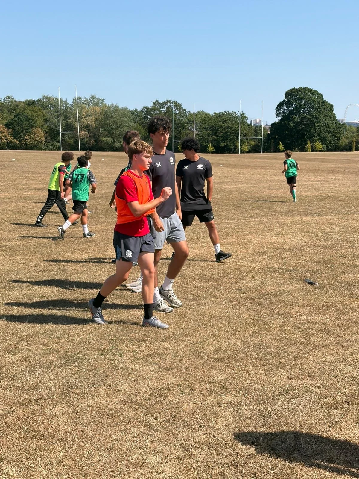 Kids and coaches training on a dry grass rugby field on a sunny day, with goal posts and trees in the background.