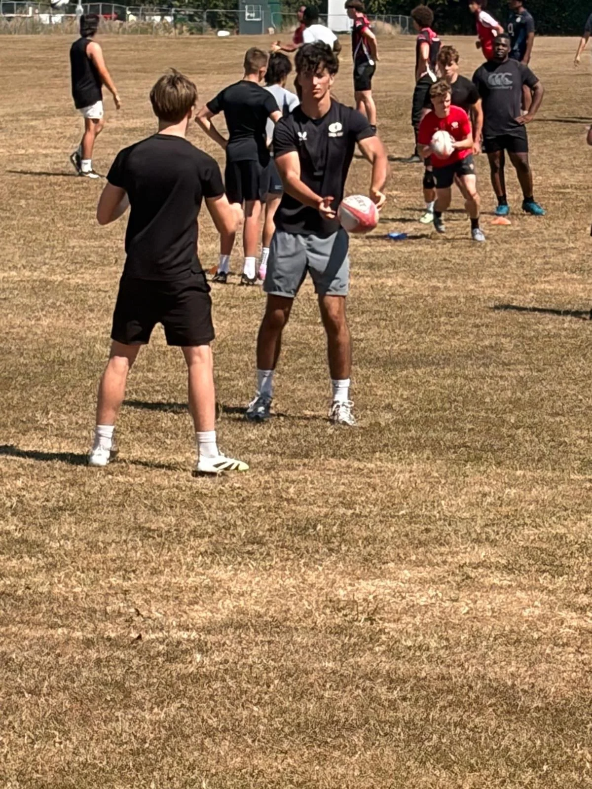 Young rugby players practicing on a field, some holding rugby balls, wearing sportswear and cleats, with a soccer goal in the background.