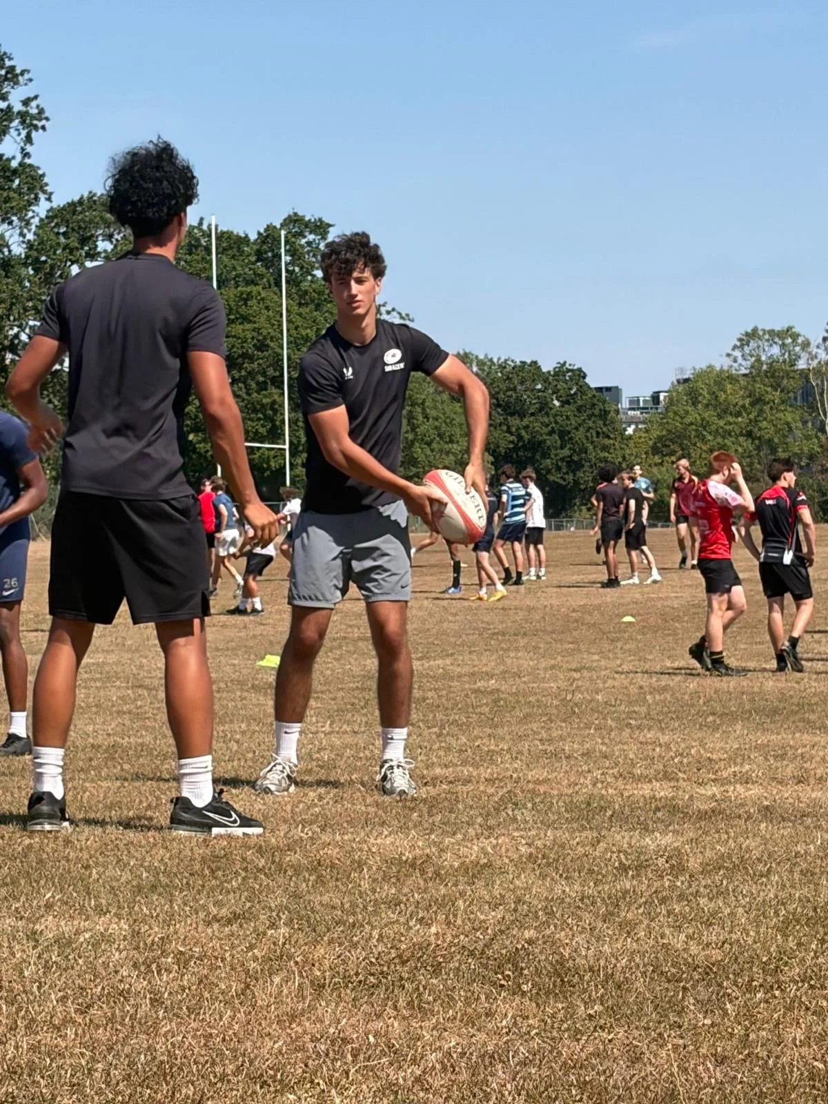 Group of young rugby players practicing on a field with two main players in the foreground, one holding a rugby ball and the other standing nearby. Other players are scattered in the background, engaging in drills or warmups on a sunny day.