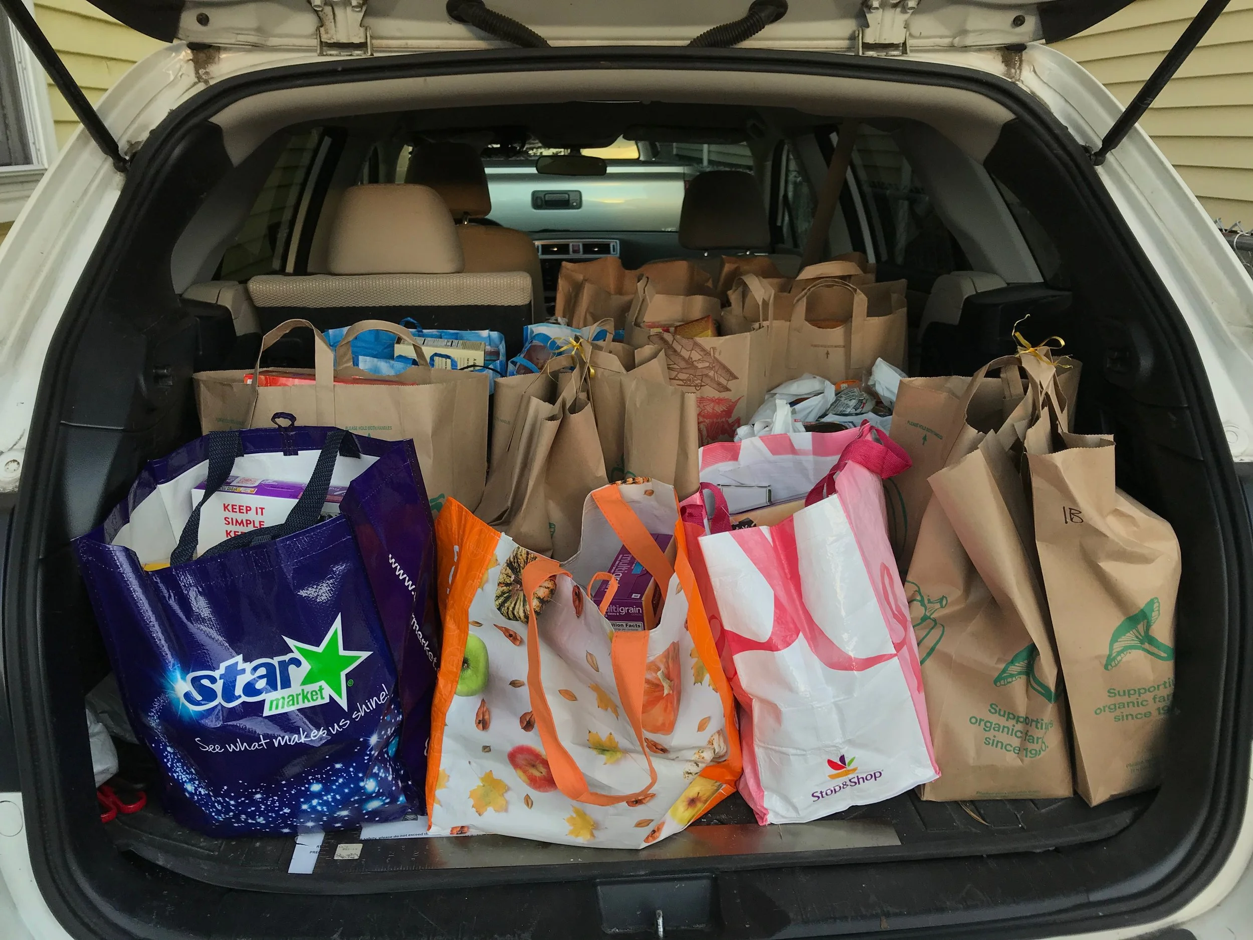 Open trunk of a car filled with paper bags of groceries, including a blue bag from Star Market, a plaid orange and white bag, and green and beige bags from Stop & Shop.