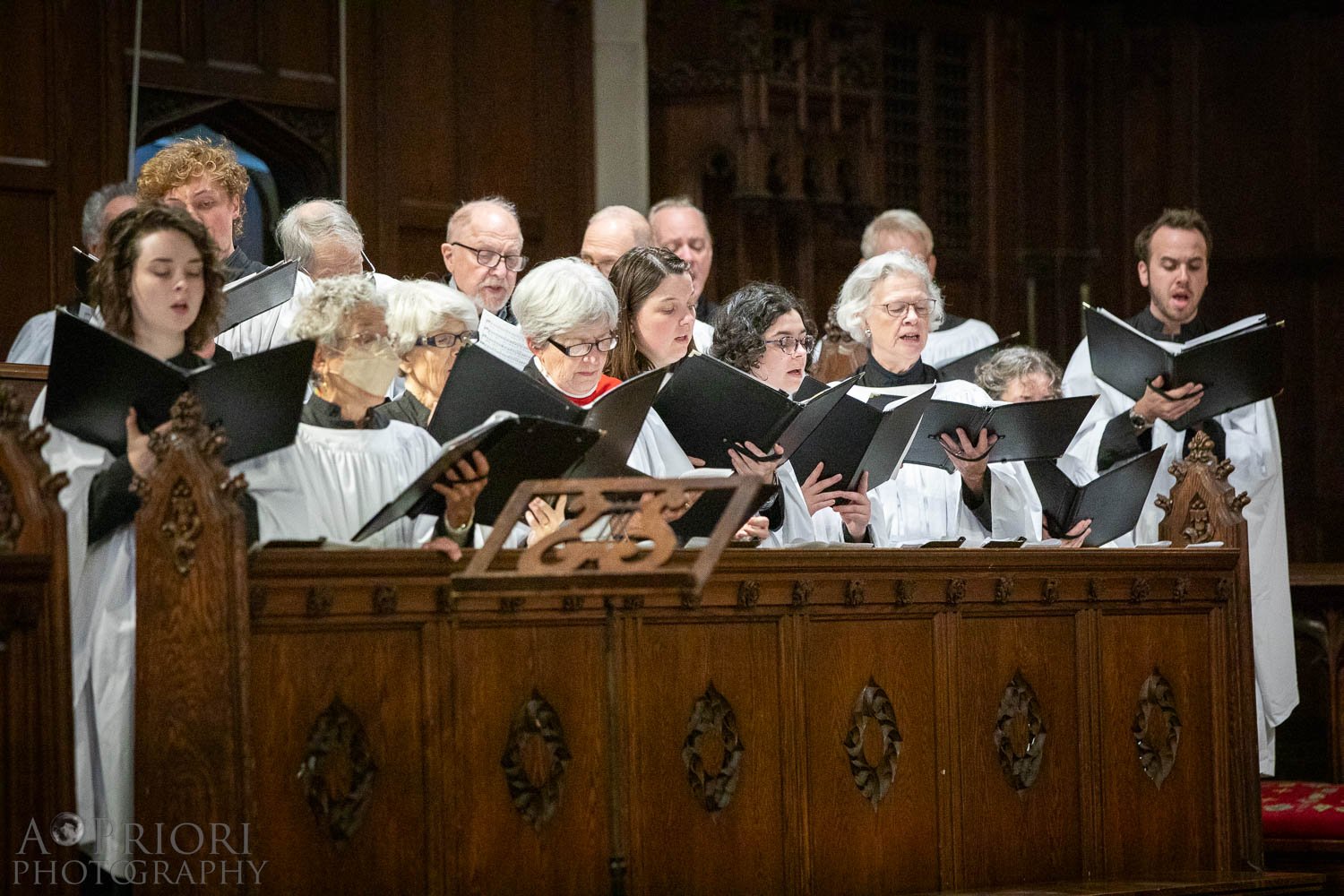 A choir of adults, dressed in white robes with black accents, singing in a church or cathedral, holding black folders with sheet music. They are standing behind a wooden choir stall with intricate carvings.