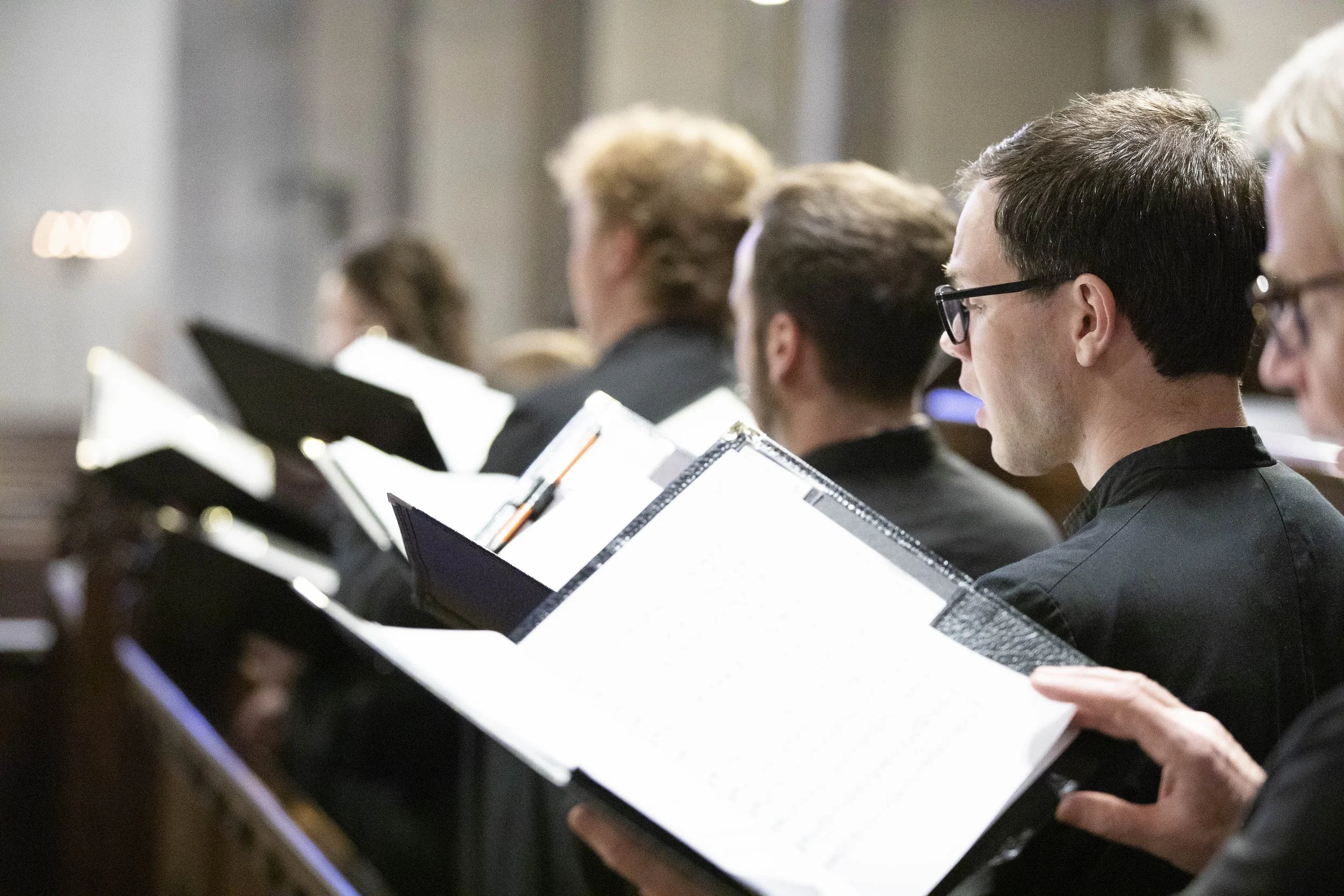 People sitting with sheet music in front of them, likely during a choir or orchestra rehearsal or performance.