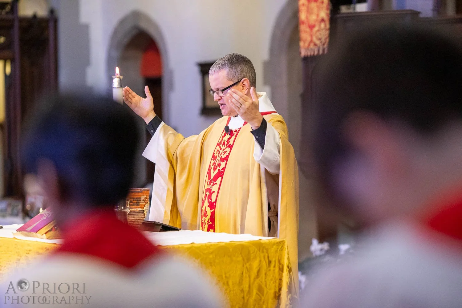 A priest wearing gold and red liturgical vestments is seen with eyes closed and arms raised during a church service inside a church with stone arches. The altar is covered with a yellow cloth.