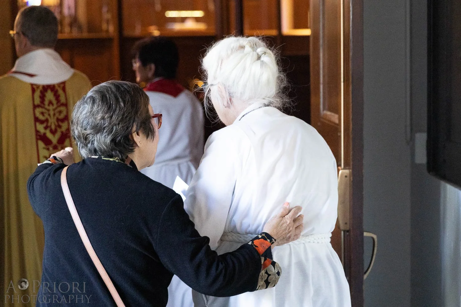 Two women, one elderly with white hair in a braid wearing a white robe, and the other middle-aged with dark hair and glasses wearing a black outfit, are in a church, talking or praying together.