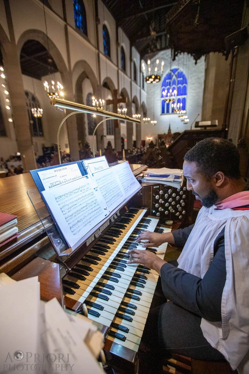 A man dressed in choir robes playing the organ inside a church. The church has high arched windows, chandelier lighting, and stained glass windows in the background.
