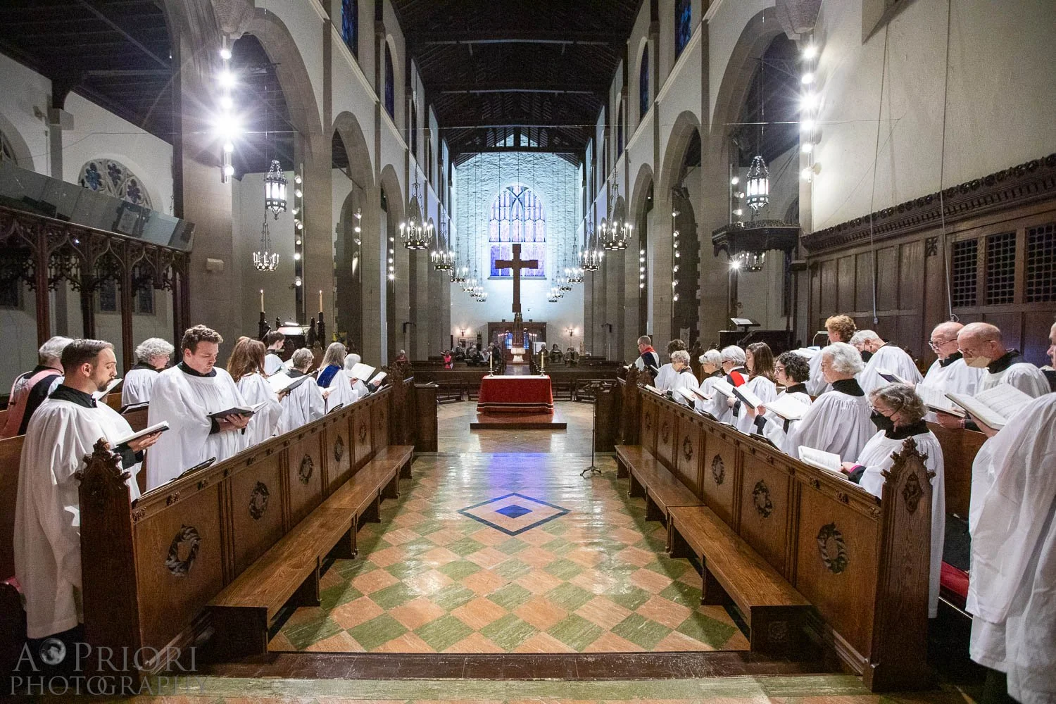 A choir dressed in white robes standing in a church nave, singing or praying, facing an altar with a cross and candles. The church has high ceilings, stained glass windows, and wooden pews.