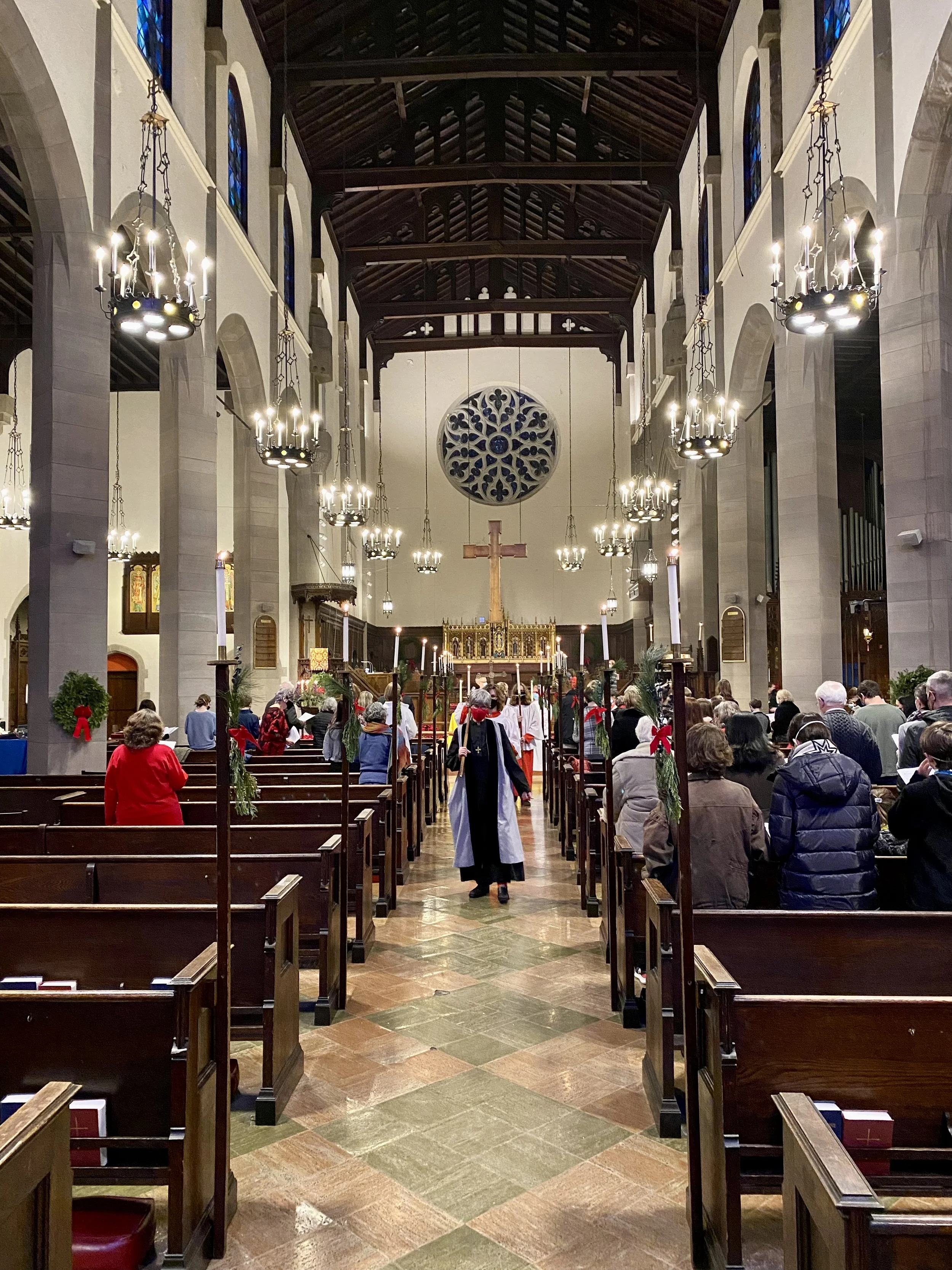 Inside a church with many rows of wooden pews, a central aisle, hanging chandeliers, stained glass windows, a large wooden cross at the altar, and people seated or standing, engaged in a religious ceremony or service.