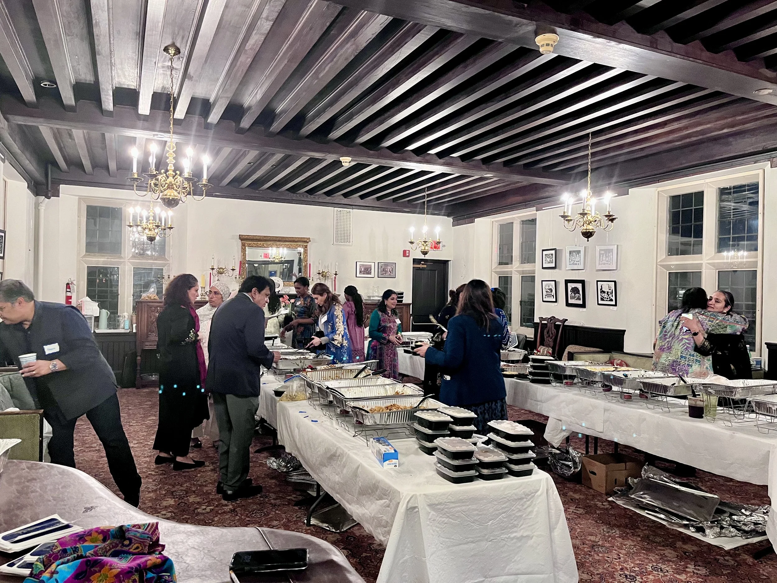 People serving themselves food at a buffet inside a dining room with white walls, framed pictures, large windows, and dark wood ceiling beams.