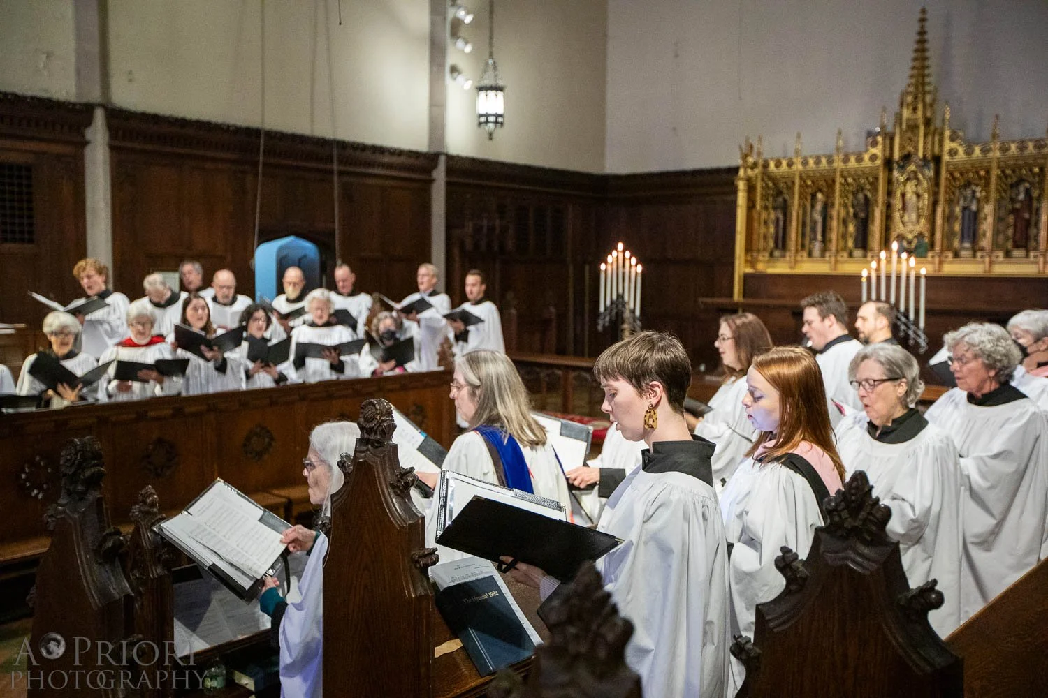 A choir dressed in white robes singing in a church with dark wooden walls, candles, and gold decorations.