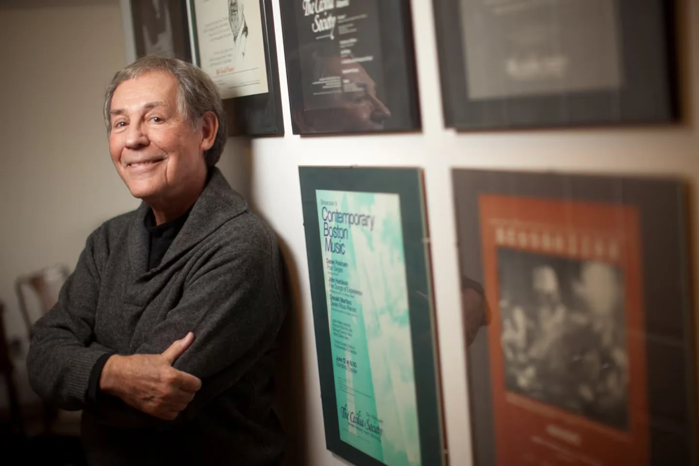 An man with gray hair and a smile, wearing a gray cardigan, leaning against a wall with framed posters or pictures.