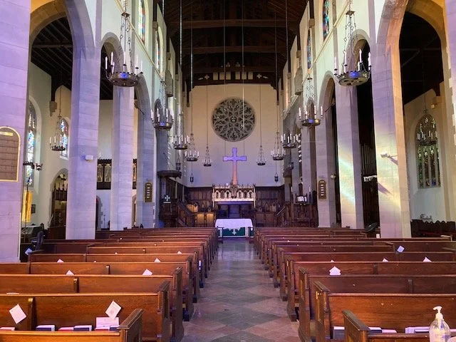 Interior of a church with wooden pews, tall columns, hanging chandeliers, and a cross at the front altar.