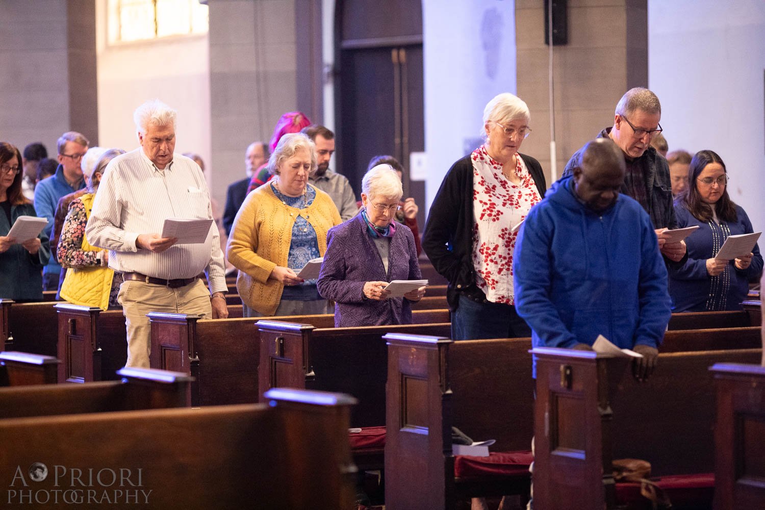 A group of people standing in pews inside a church, holding hymn books or prayer sheets, engaged in prayer or hymn singing.