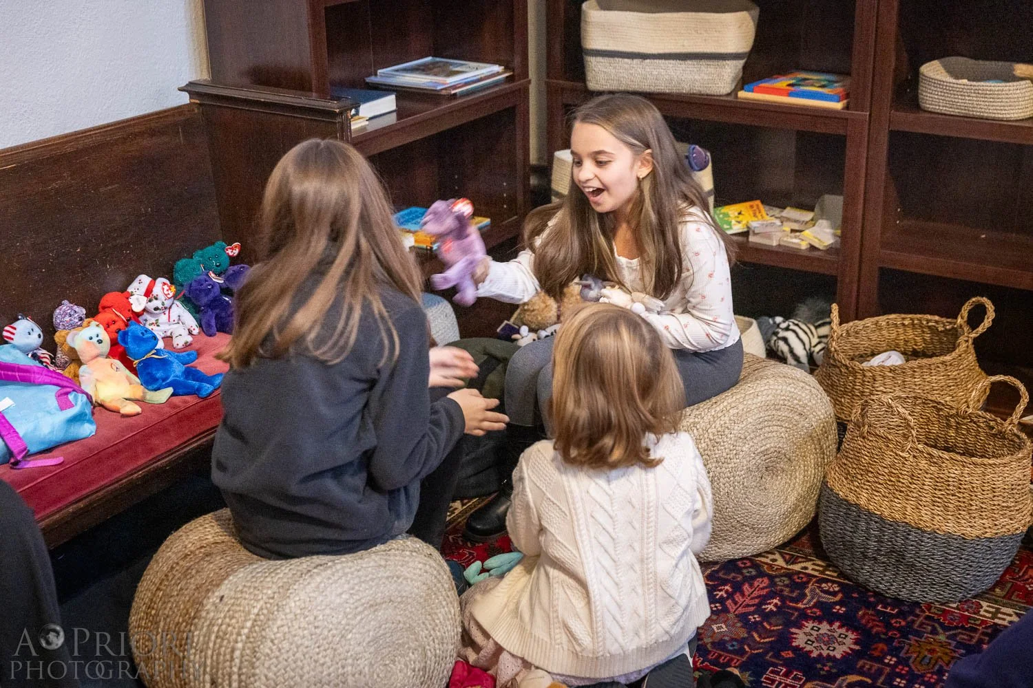 Four girls sitting and standing around in a cozy room with wooden shelves and baskets. They are engaged in a playful interaction, exchanging plush toys, with a collection of plush toys visible on a nearby bench.