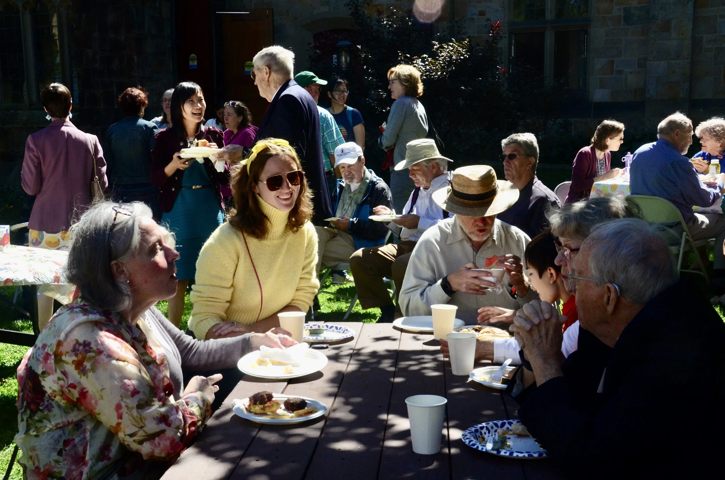 A group of people gathering outdoors, sitting at tables with food and drinks, enjoying a sunny meal or celebration in a garden.