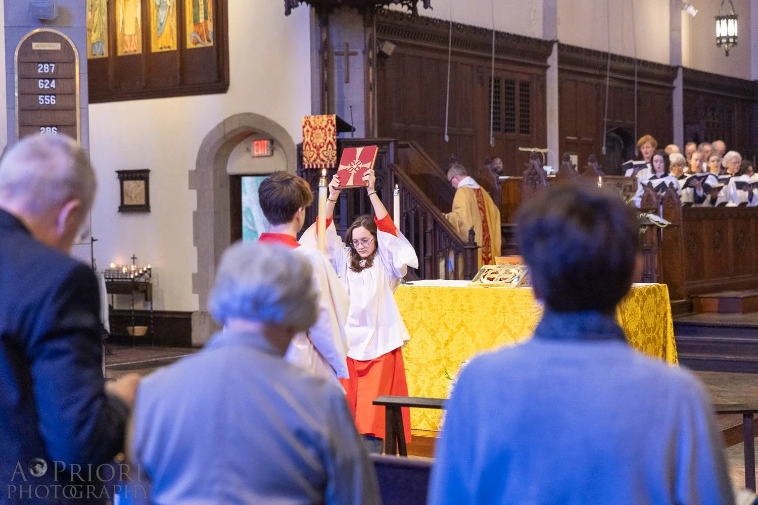 A girl dressed in white and red robes holding a red book with a white cross above her head during a church service, with a congregation and choir in the background.