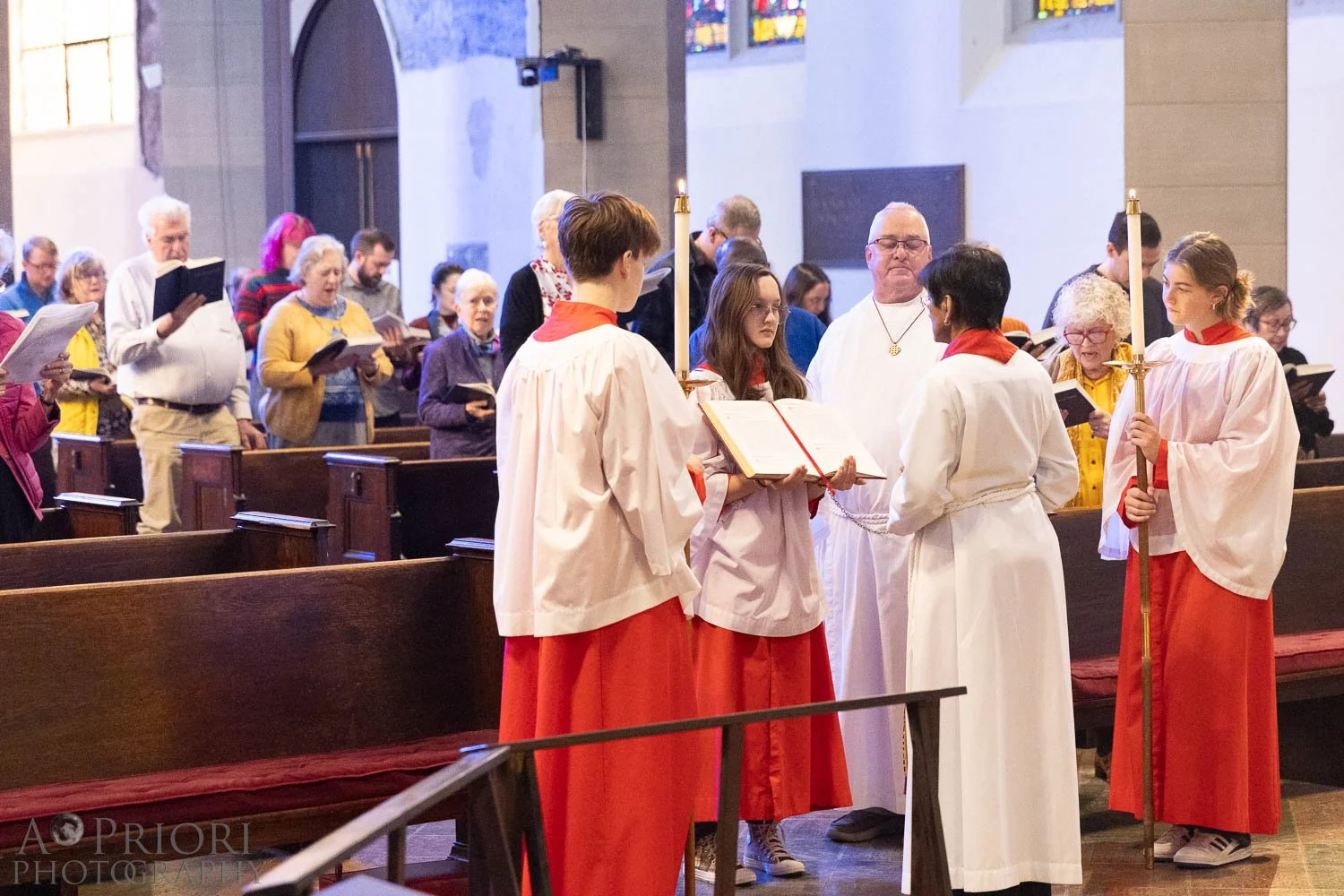 People gathered in a church during a religious ceremony, some are reading from hymnals or prayer books, and altar servers dressed in white and red robes holding candles and a cross.