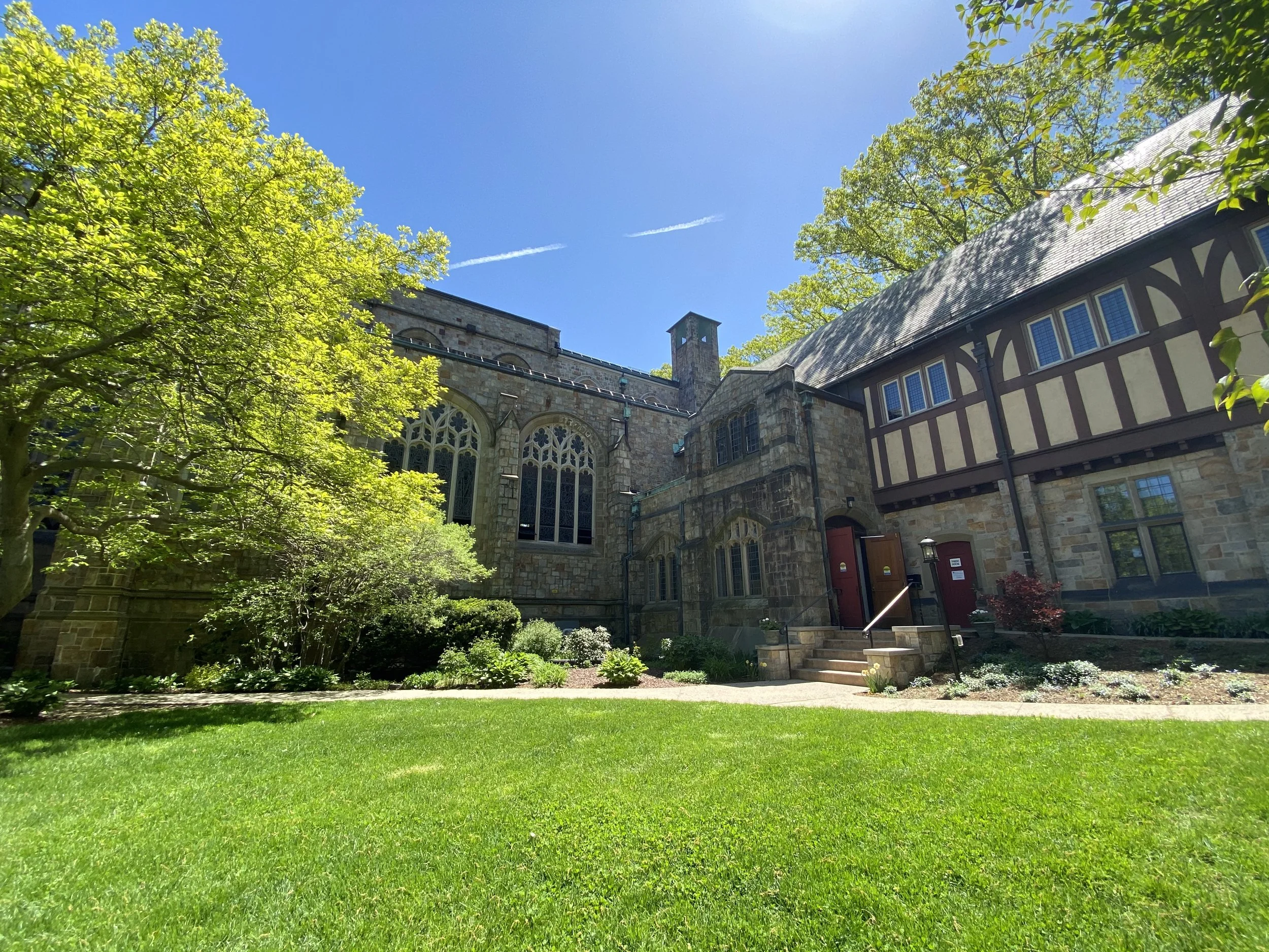 View of a historic stone building with large windows, surrounded by trees and a well-maintained green lawn under a clear blue sky.