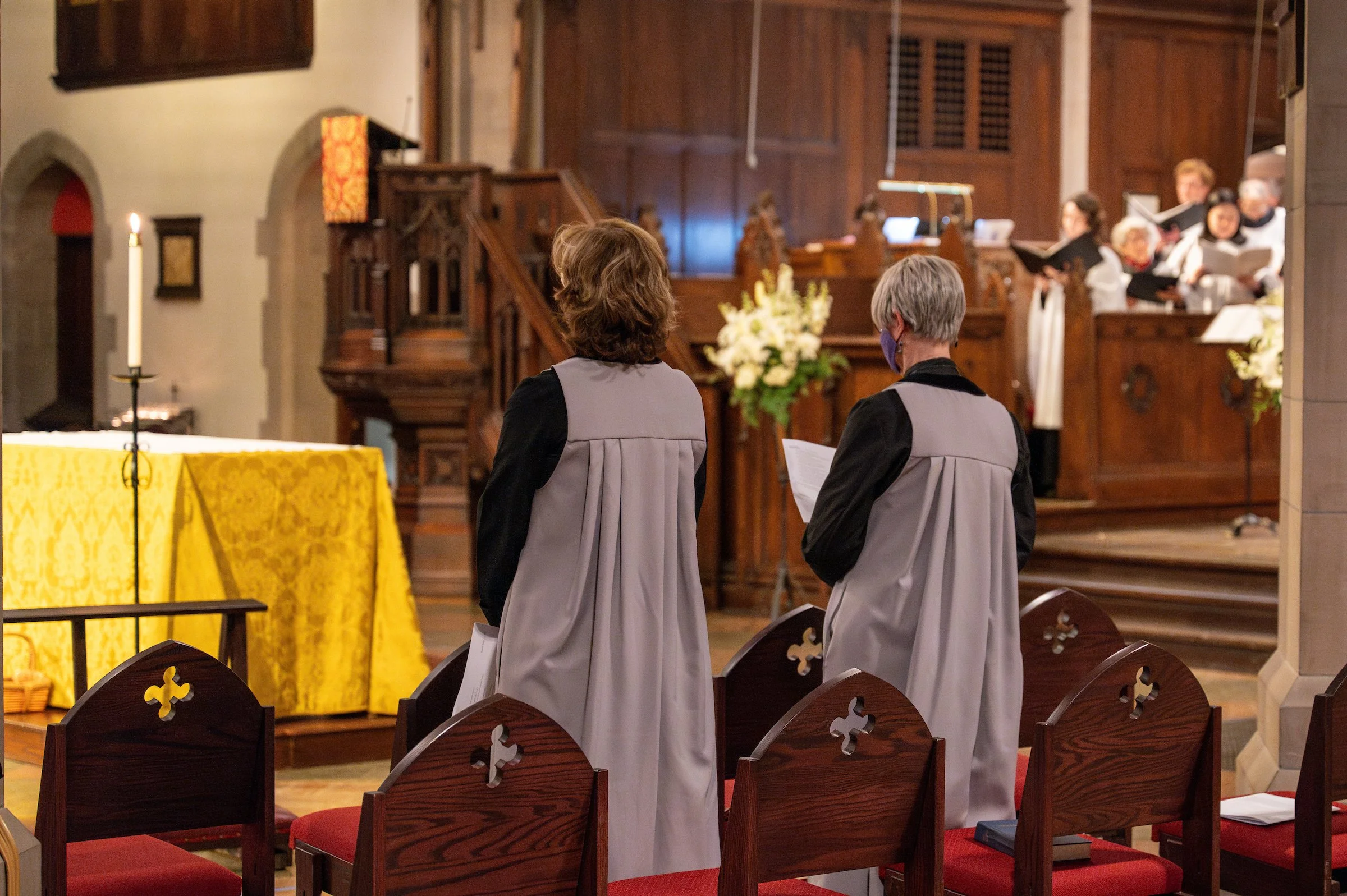 Two women wearing choir robes stand in a church during a service, facing the altar with their backs to the camera. In the background, a choir is singing in front of a wooden altar with flowers, and a large candle is on a stand near the altar.