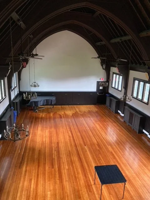 Empty event hall with polished wooden floor, black stage at the front, and tables on the sides, illuminated by natural light from multiple windows.