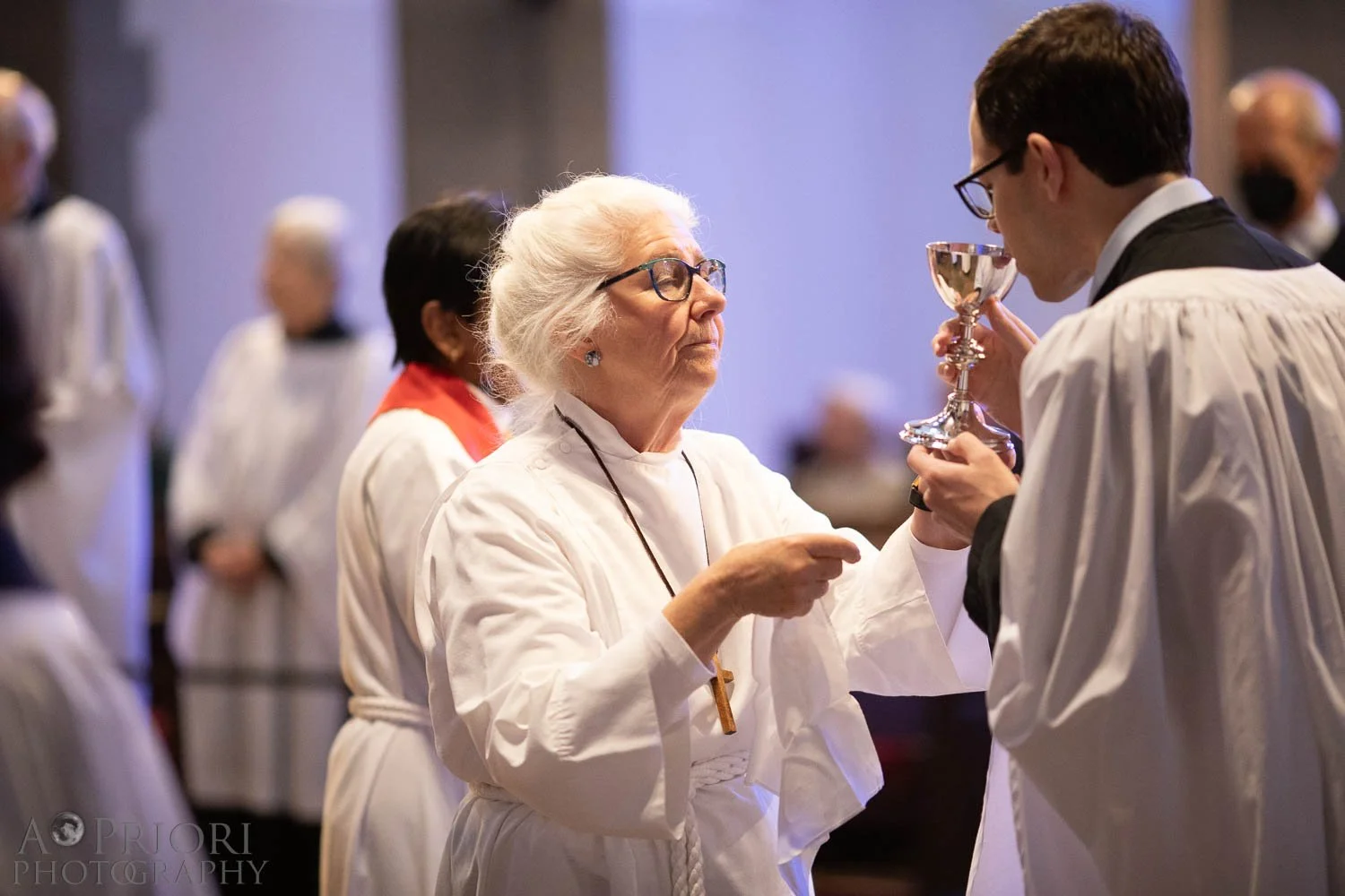 An elderly woman, wearing glasses and a white clergy robe, points at a chalice held by a young clergy man in the church choir. The young man is wearing a clerical robe and glasses, and is drinking from the chalice. Other altar servers and clergy are in the background.
