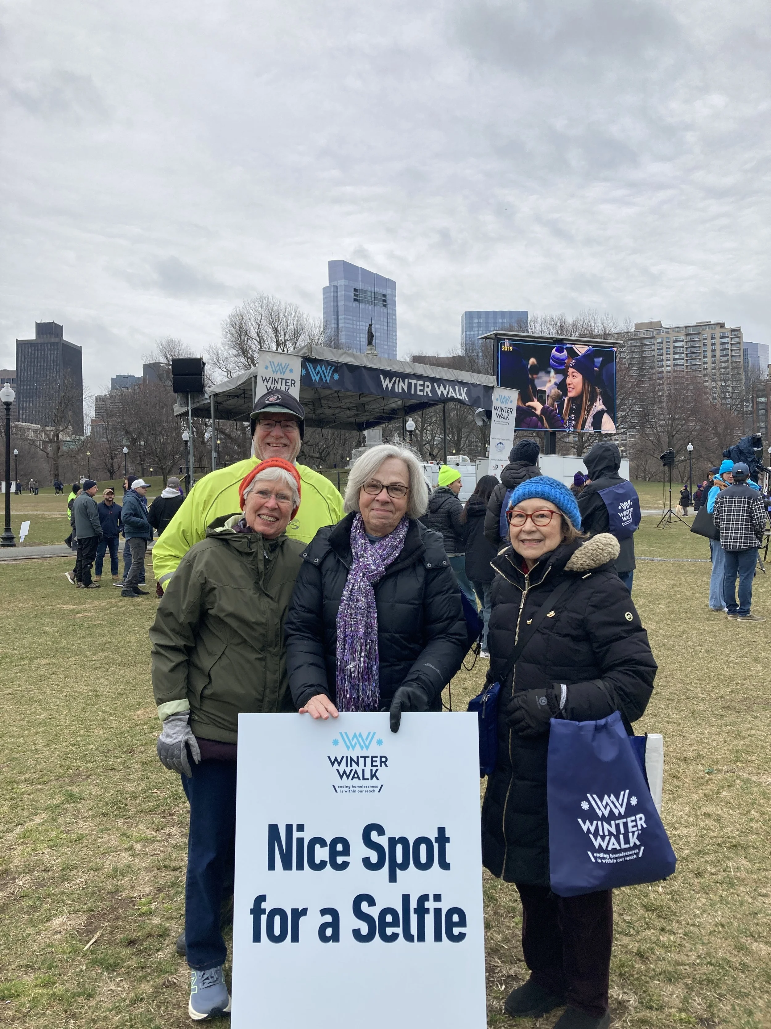 Four people smiling at a winter walk event holding a sign that says, 'Nice Spot for a Selfie.' They are outdoors in a park with tall buildings in the background, wearing warm clothing and winter hats.