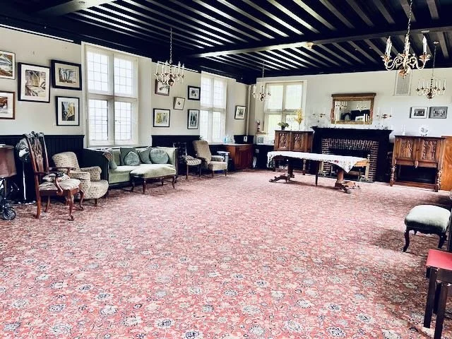 Spacious vintage living room with antique furniture, framed artwork on white walls, large windows, and a brick fireplace with a mirror above.