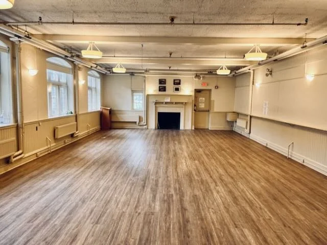 Empty event hall with hardwood floor, large windows, and a fireplace on the back wall.