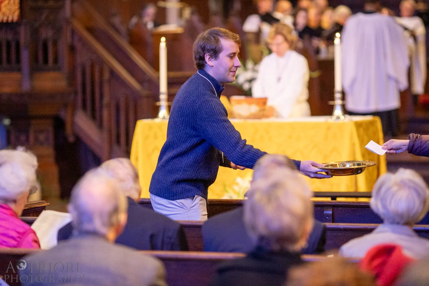 A young man in a navy sweater is placing a donation in a silver tray during a church service or ceremony, with elderly people seated in pews and a table with candles and a yellow cloth in the background.