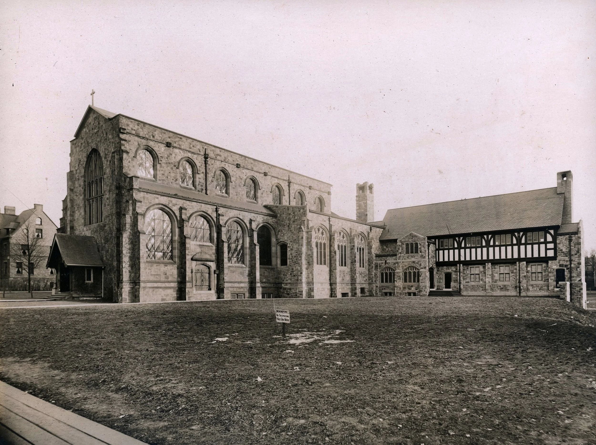 Black and white photograph of an old stone church with large arched windows and a steeple with a cross on top, surrounded by a barren dirt area.