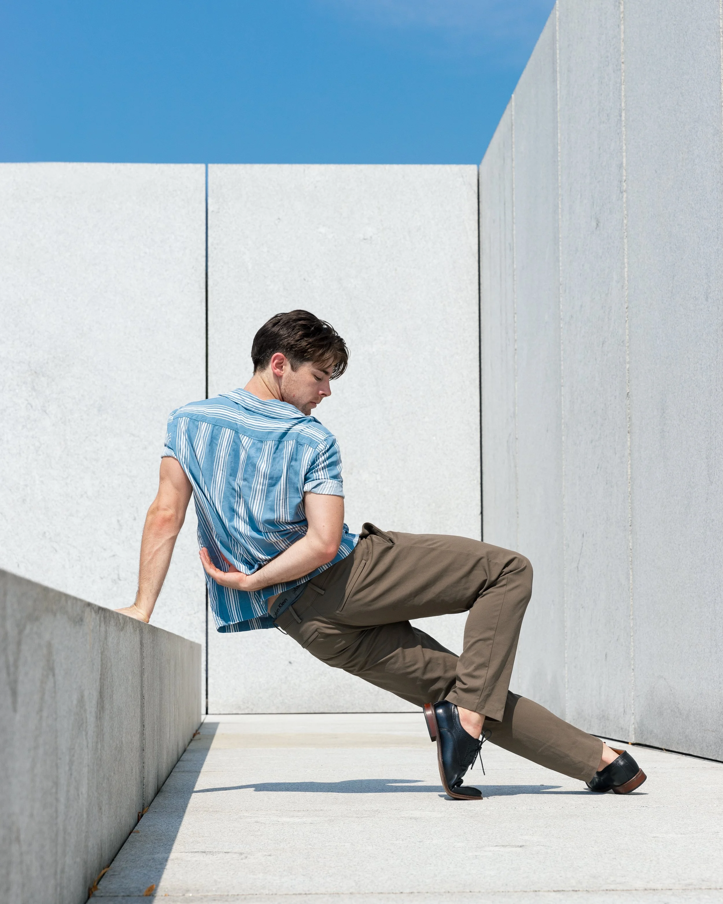 Young man in a striped shirt and khaki pants sitting on a concrete ledge with his head down and one hand on his side, against a modern concrete wall and bright blue sky.
