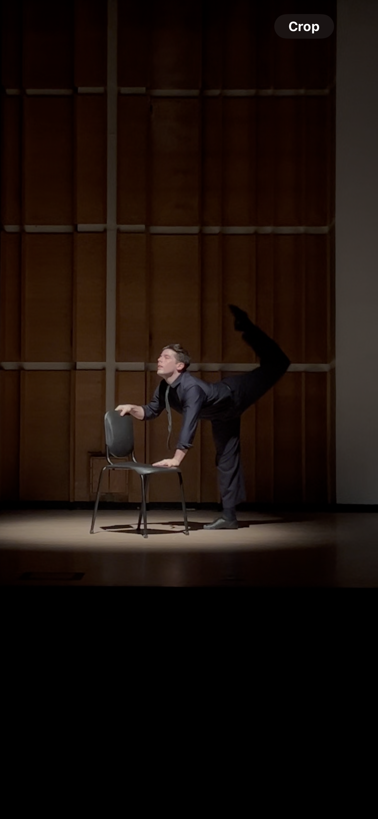 A man in formal attire performing a pose on stage, with one hand on a chair and one leg raised behind him, against a wooden wall background.