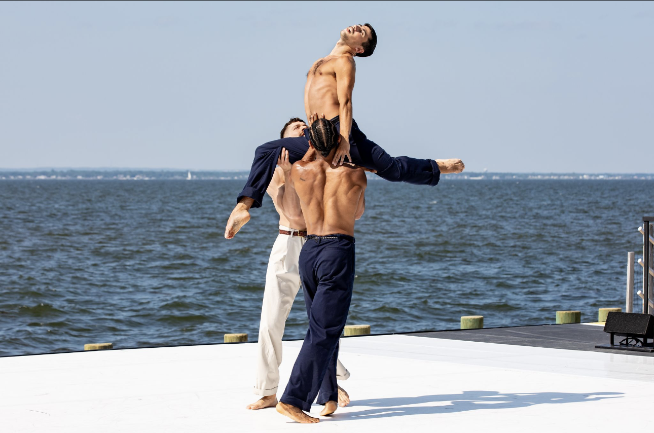 Three shirtless men perform a martial arts routine on a white platform by the water, with one man holding another in a hoop while the third supports him from below.
