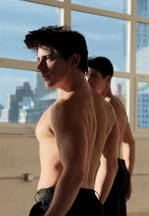 Two shirtless men with muscular physiques standing in a gym, with windows and city buildings visible in the background.