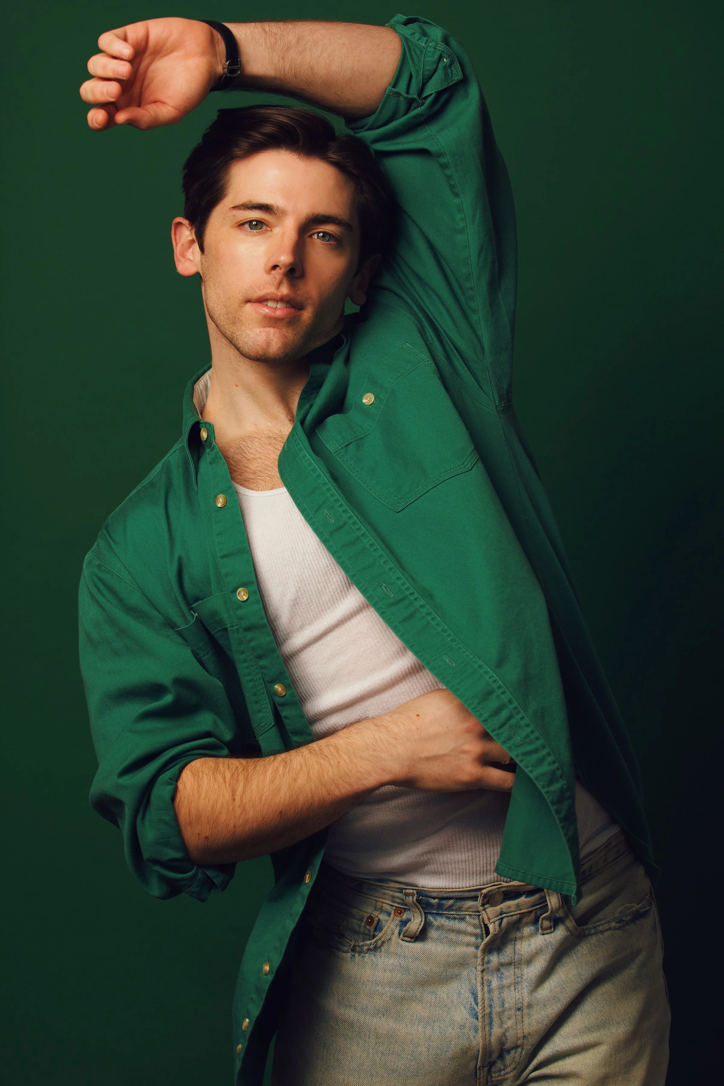 A young man with dark hair wearing a green shirt over a white t-shirt and jeans, posed with one arm raised above his head and the other hand resting on his stomach, against a green background.
