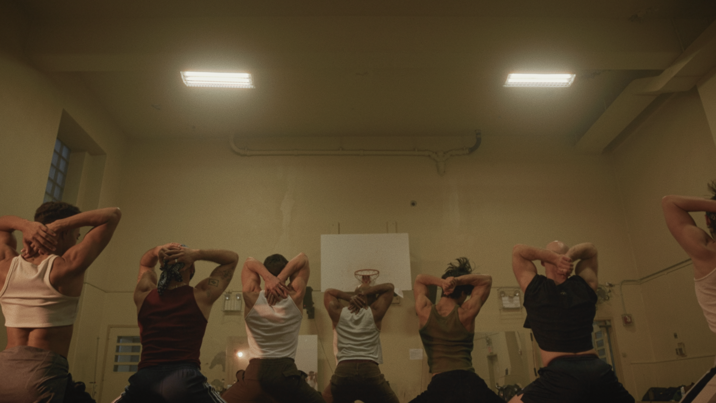A group of people participating in a yoga or fitness class with their hands behind their heads, in a room with fluorescent ceiling lights and a basketball hoop on the wall.