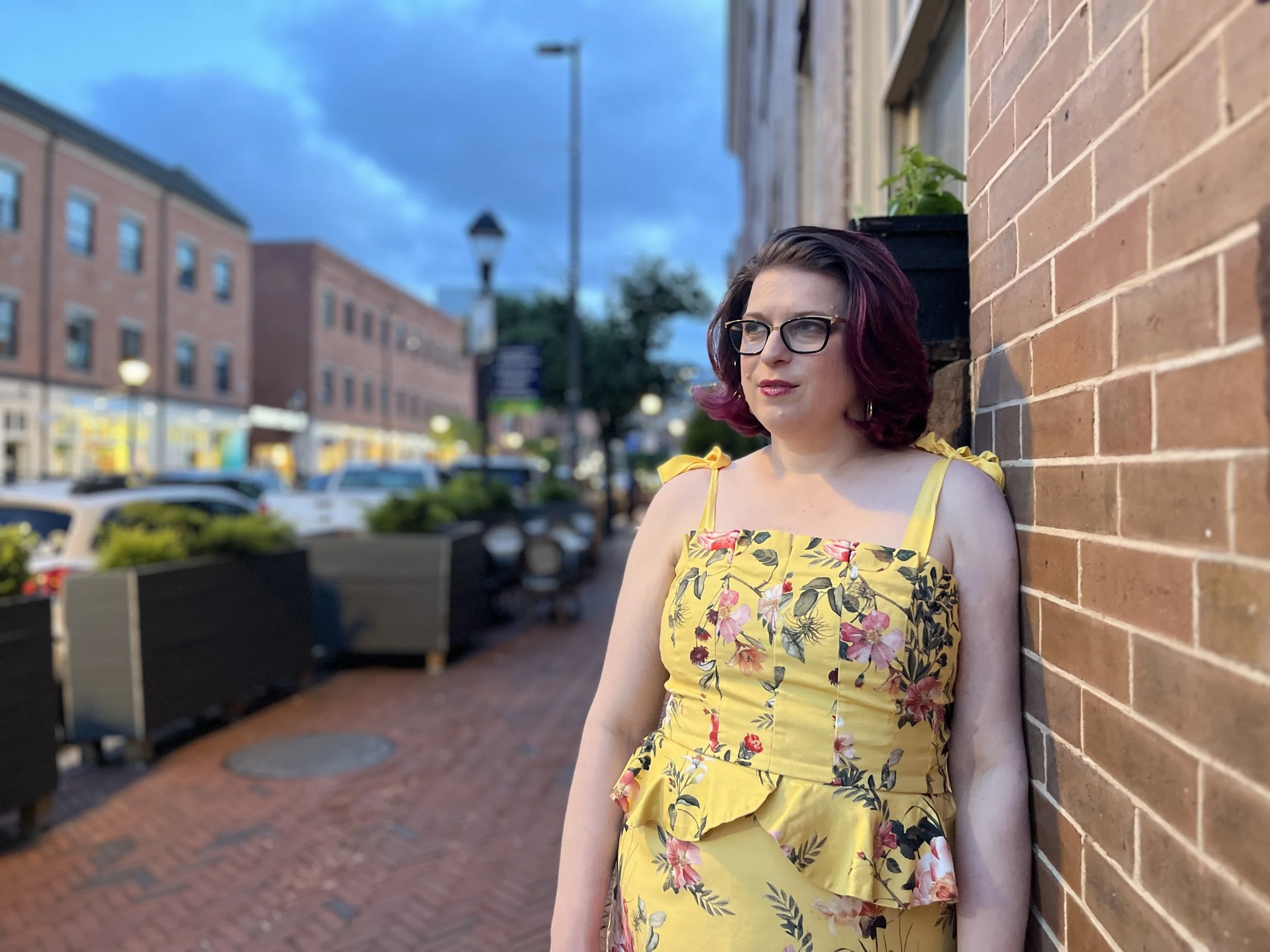 A woman with dark hair and glasses, wearing a yellow floral dress, leaning against a brick wall on a city street during the evening or twilight.