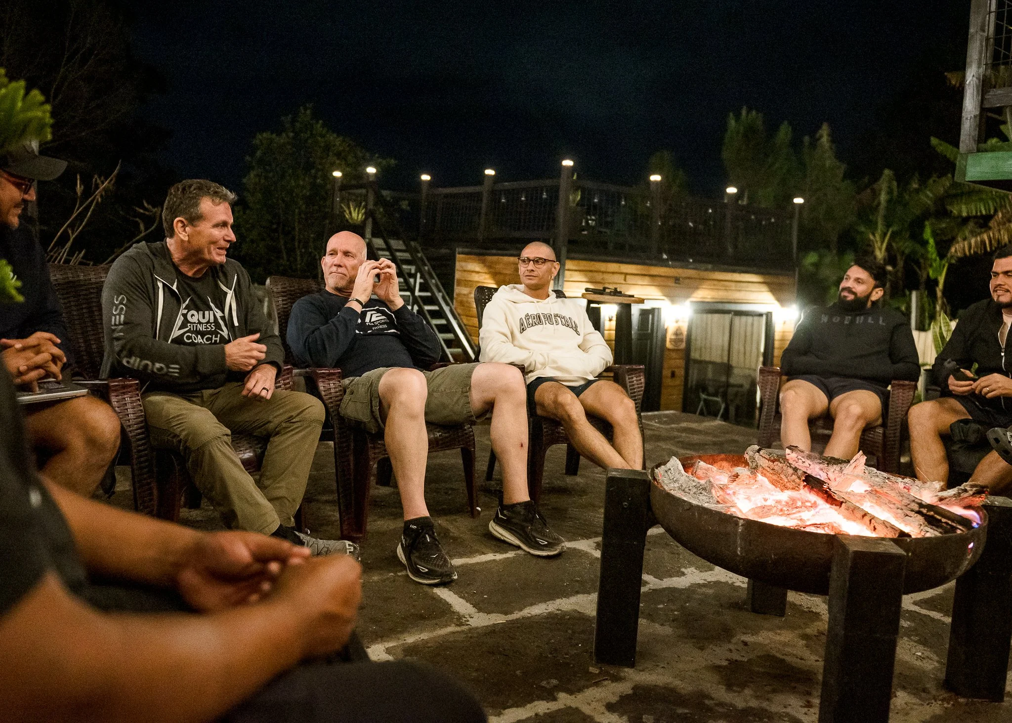 Group of men sitting outdoors at night around a fire pit, some talking and some listening, with trees and a building in the background.