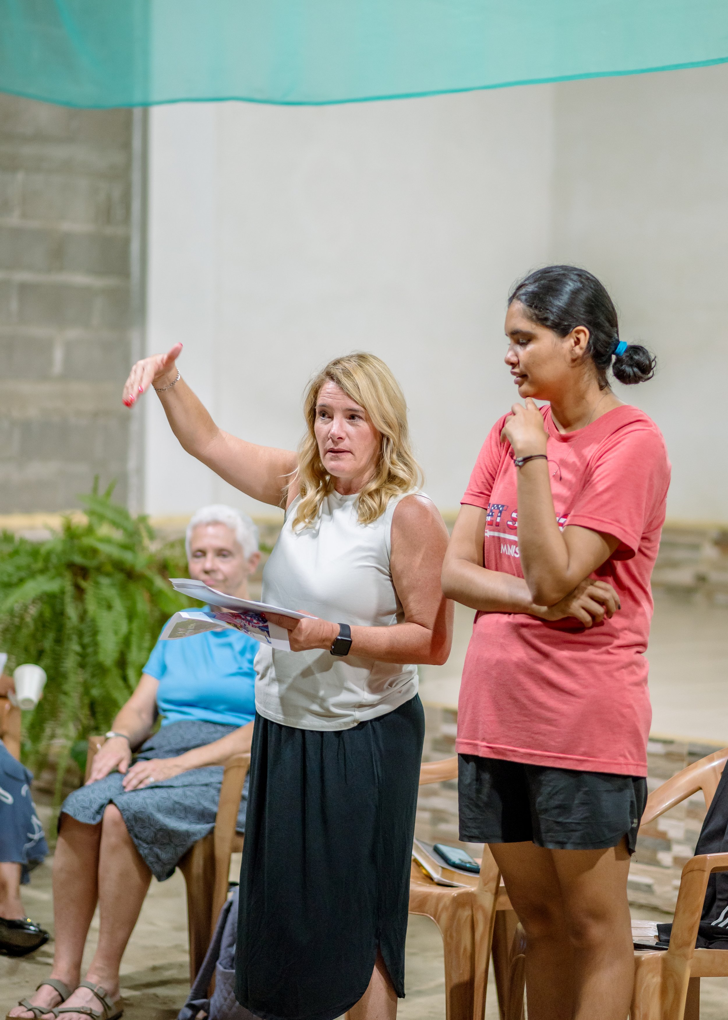 A group of people in a room, with two women standing and talking, one with a hand raised gesturing while the other listens. Several chairs and a plant are visible.