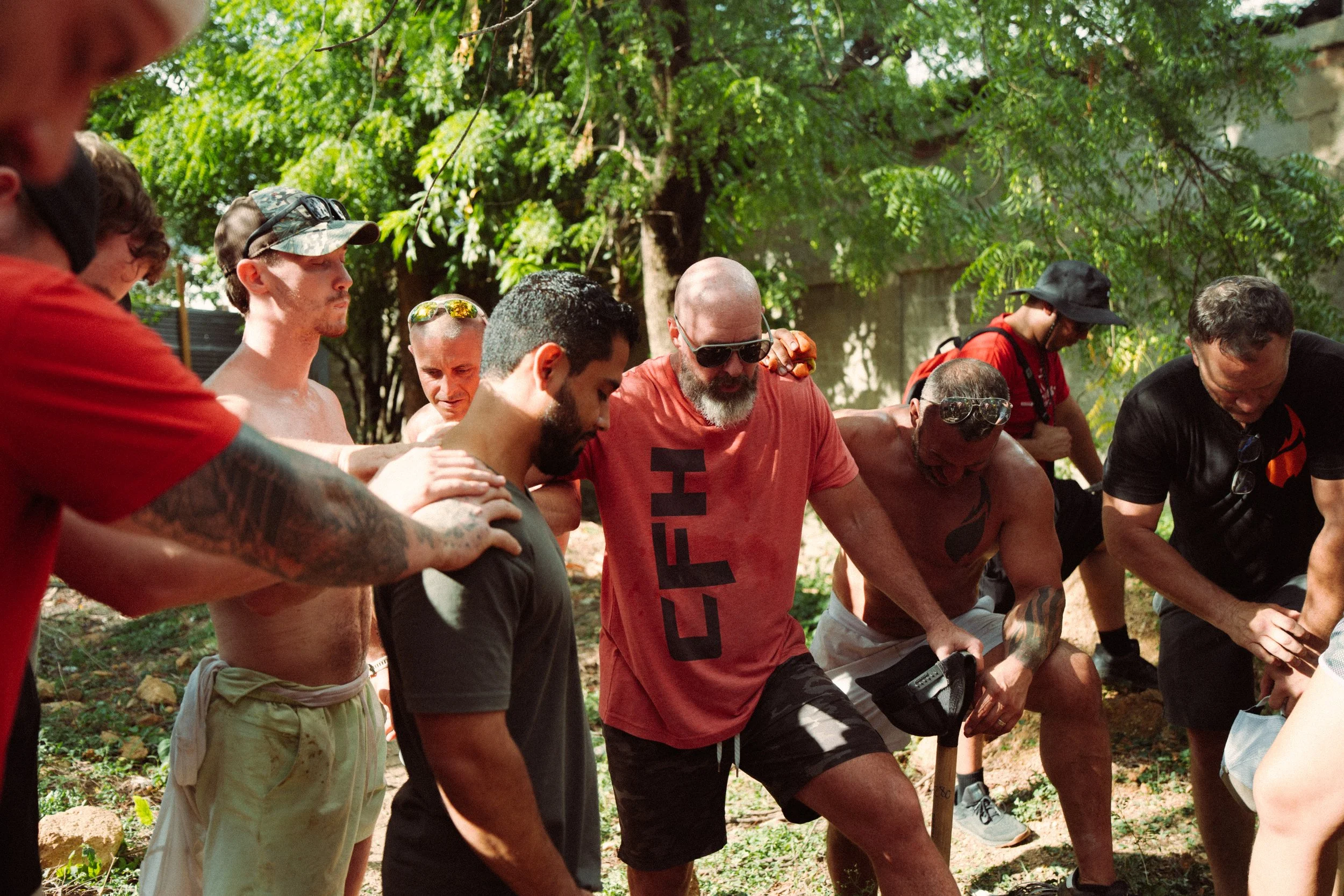 A group of diverse men gathered outdoors in a circle, praying or meditating with their eyes closed, surrounded by trees and greenery.