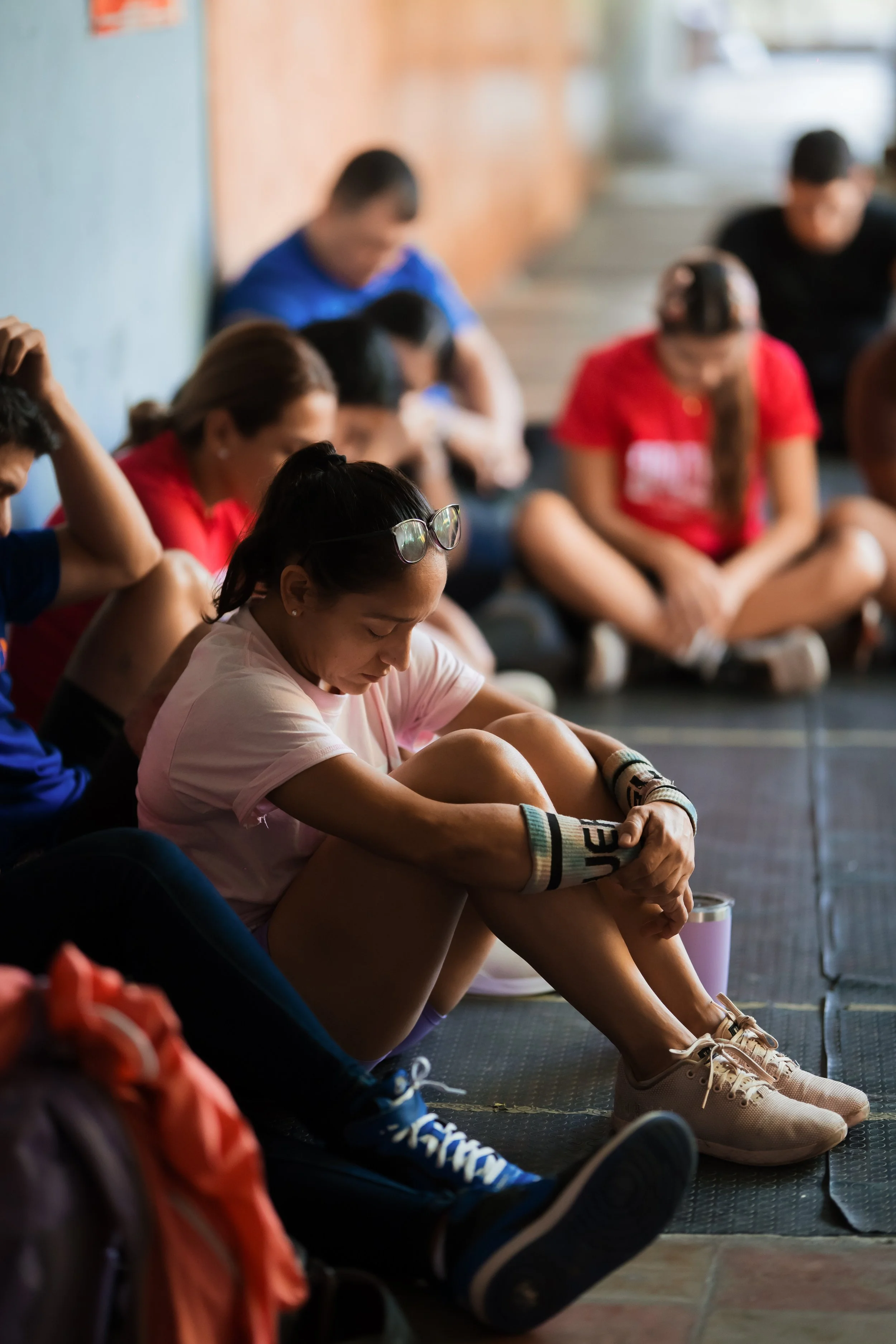 Group of young people sitting on the floor with their heads bowed and eyes closed, appearing to meditate or pray in a room with wooden walls.