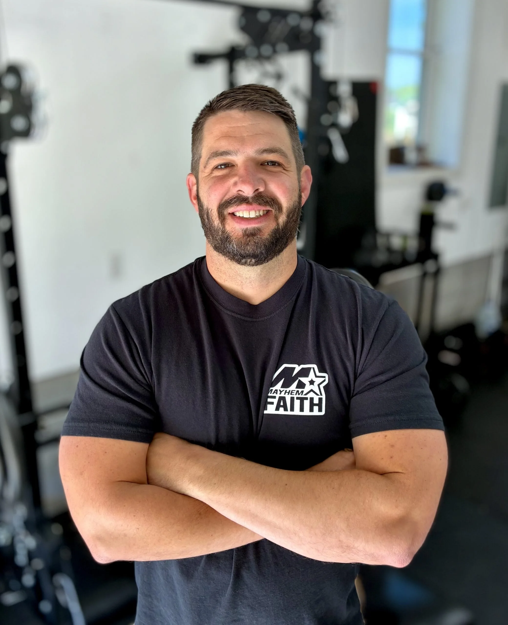 A man with short brown hair and a beard standing with arms crossed, smiling, wearing a black T-shirt with text and logo, in a room with gym equipment.