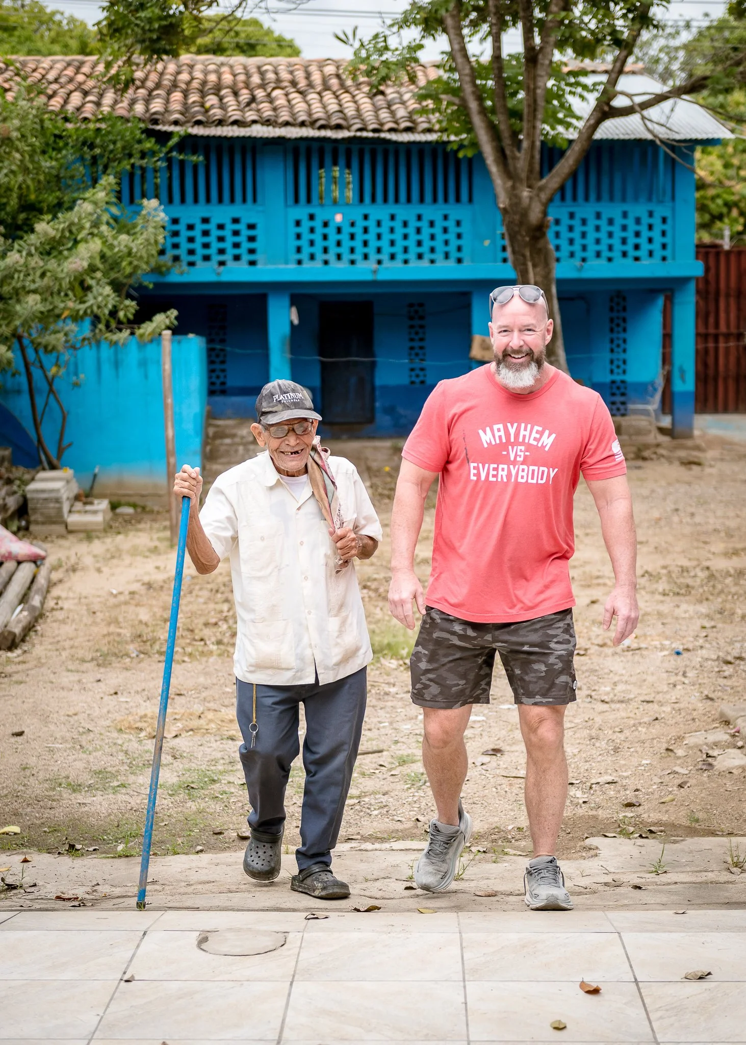 An elderly man and a younger man walking together outdoors. The elderly man is holding a cane and wearing a cap, glasses, a light shirt, and dark pants. The younger man has a beard, sunglasses on his head, a red t-shirt with the phrase 'Mayhem vs Everybody', and camouflage shorts. They are smiling and walking on a dirt path in a backyard with a blue house and trees in the background.