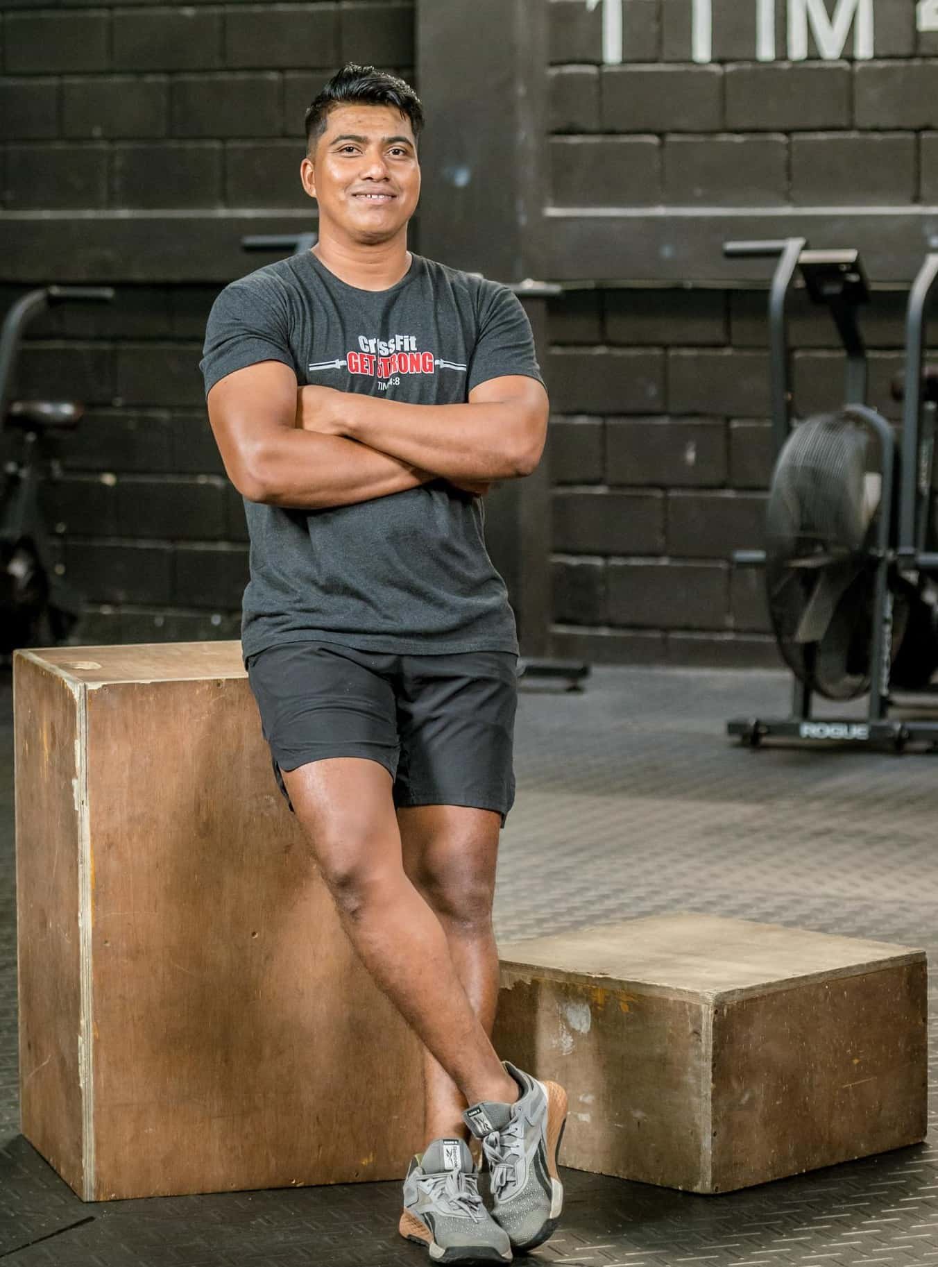 A man standing with arms crossed and leaning against a wooden box in a gym. He is wearing a dark grey t-shirt, black shorts, and grey running shoes. The background shows gym equipment and a black wall with large white lettering.